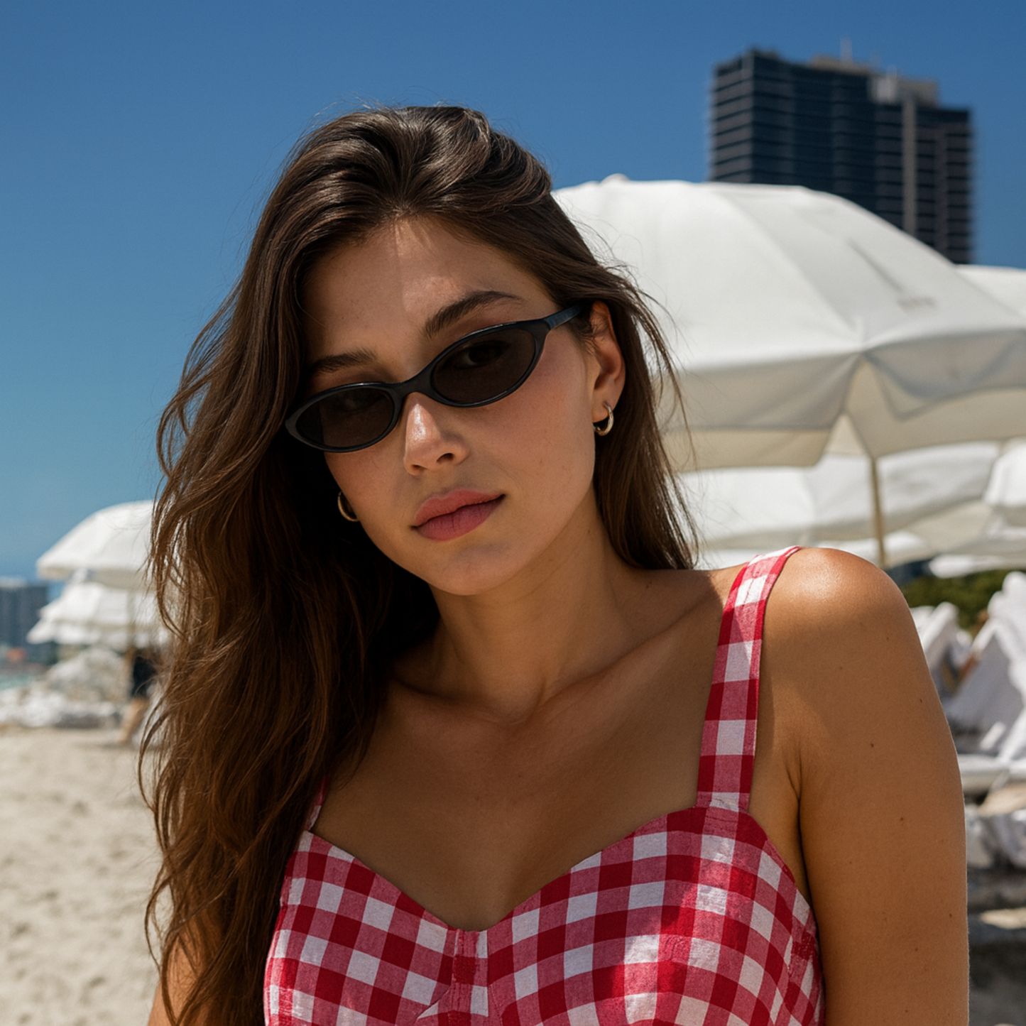 Woman wearing sunglasses and a red checkered dress on a beach with white umbrellas and buildings in the background.